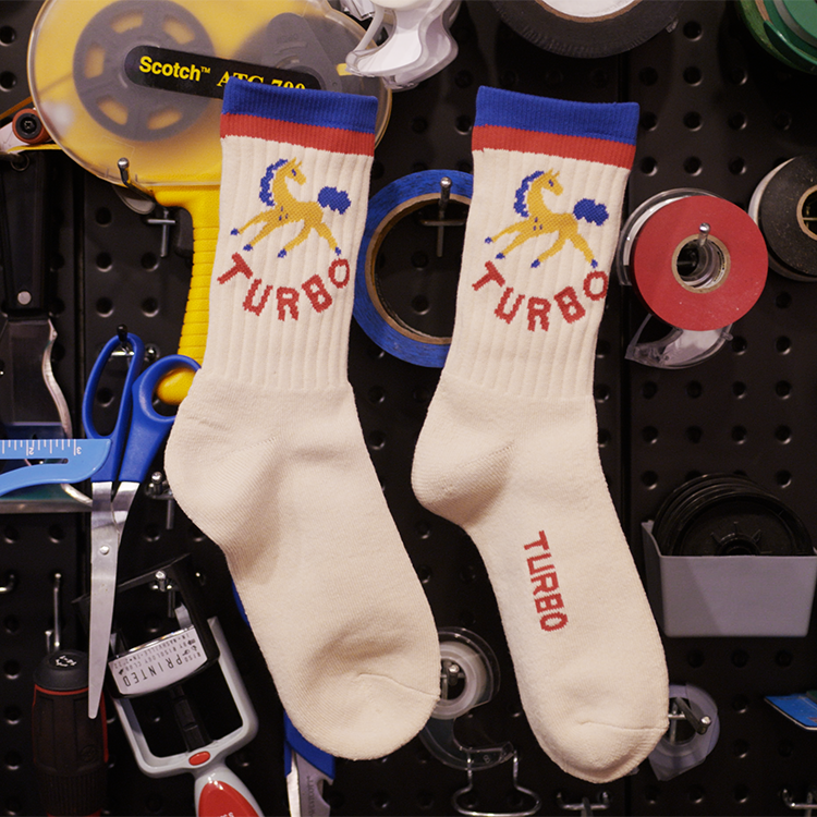 A pair of off-white crew socks hanging on a black pegboard filled with tools. The socks feature a blue and red striped cuff, a yellow horse graphic, and the word 'TURBO' in red.