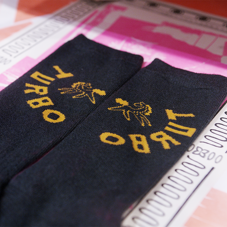 A close-up of a pair of Dark Green Merino wool socks with gold embroidered text and graphics, laid diagonally on top of a pink and white printed background.