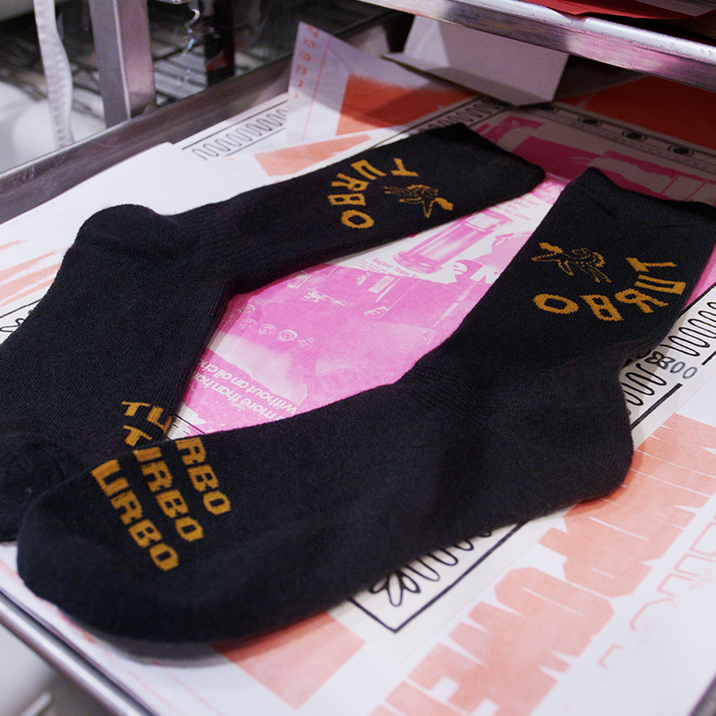 A pair of hunter green crew socks with golden 'TURBO' text and graphics, displayed on top of a pink and white printed sheet on a metal tray.